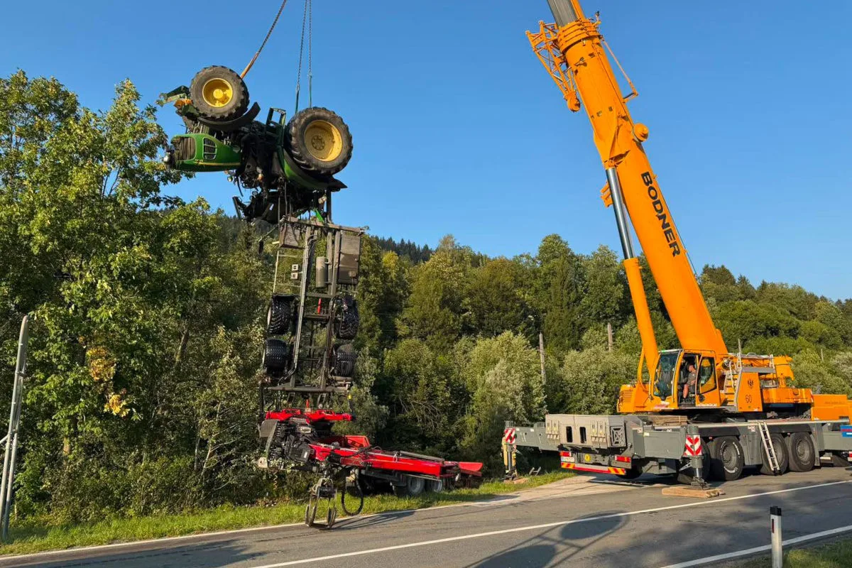 Ein großer gelber Kran hebt einen umgekippten Traktor aus einem bewaldeten Gebiet neben einer Straße. Der Traktor ist in grüner und gelber Farbe.