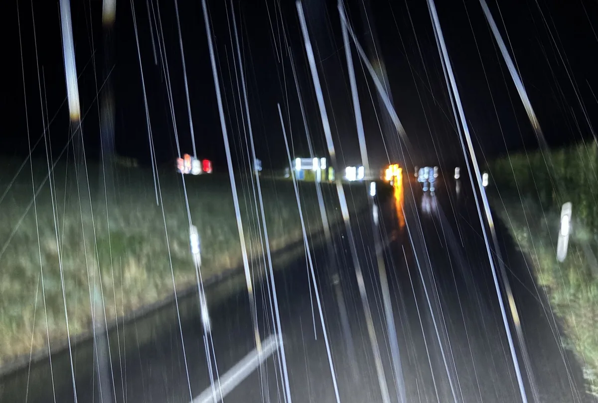 Eine stark regennasse und dunkle Straße bei Nacht ist zu sehen. Im Hintergrund sind verschwommene Lichter.