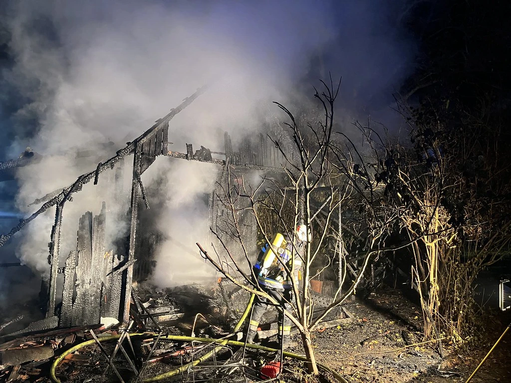 Ein Feuerwehrmann mit Atemschutz ist dabei, ein stark beschädigtes und qualmendes Gebäude bei Nacht zu löschen. Umgeben von verbrannten Strukturen und dichter Vegetation.