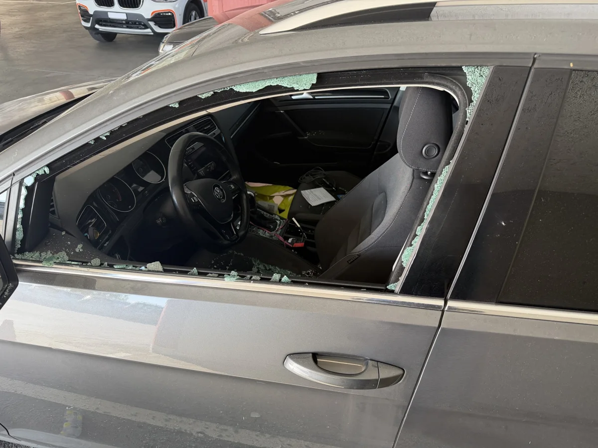 Das Bild zeigt die zerbrochene Scheibe eines grauen Autos, geparkt in einem Parkhaus. Glassplitter liegen auf dem Sitz und am Boden.
