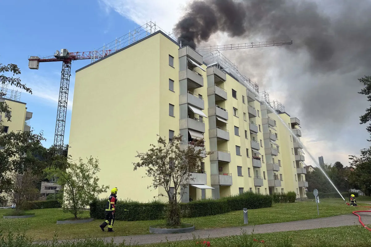 Ein Wohngebäude mit dicken Rauchwolken, die vom Dach aufsteigen. Feuerwehrleute sind unten im Einsatz mit einem Löschschlauch.