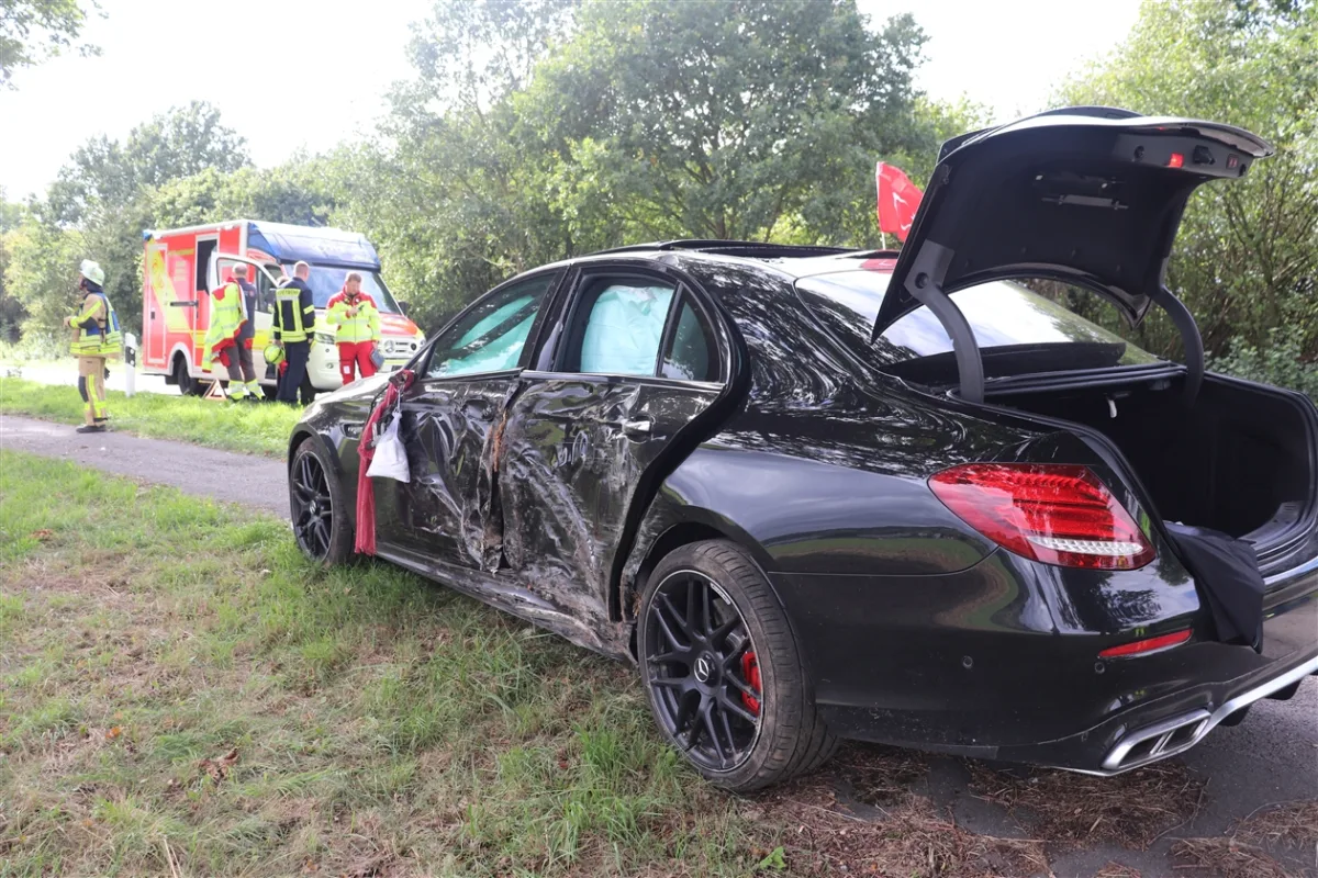 Ein schwarzer Mercedes mit schweren Seitenschäden steht am Straßenrand, während Rettungskräfte hinter dem Fahrzeug beschäftigt sind.