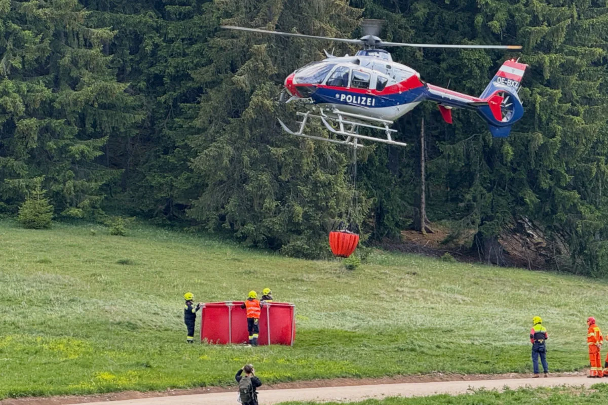 Ein hoher, zweifarbiger Polizeihubschrauber trägt einen Löschwasserbehälter über einer grünen Wiese am Waldrand. Feuerwehrleute stehen daneben.