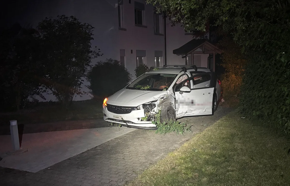 Ein weißes Auto mit erheblichem Frontschaden steht nachts auf einem Gehweg vor einem Wohngebäude. Das Fahrerfenster ist geöffnet.