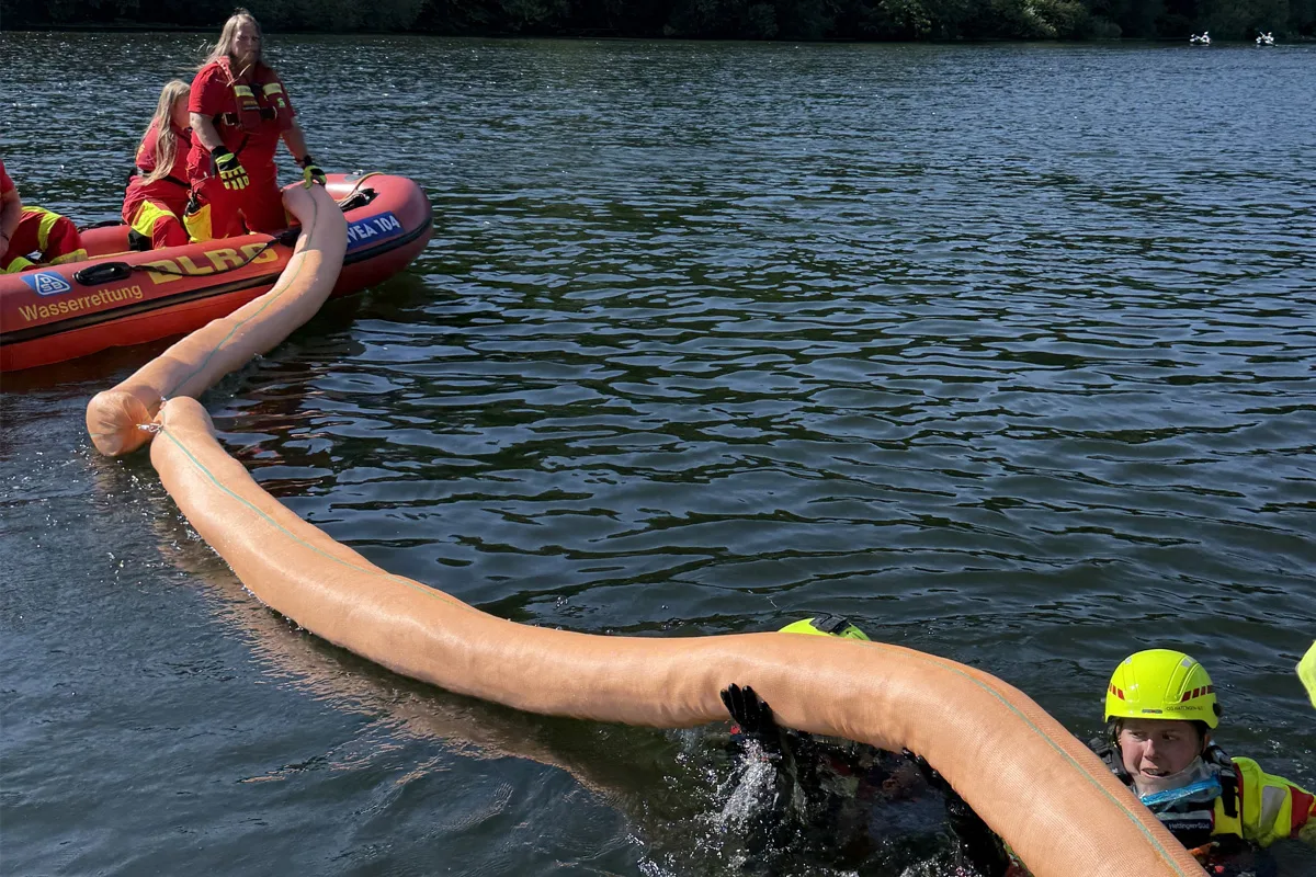 Ein DLRG-Rettungsboot mit mehreren Einsatzkräften legt eine Ölsperre auf einem See aus. Eine Person ist im Wasser.