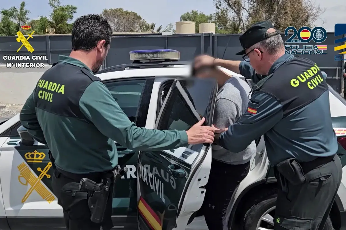 Zwei Guardia Civil-Beamte führen eine Person zu einem Fahrzeug. Logos der Guardia Civil und Policia Nacional sind sichtbar.
