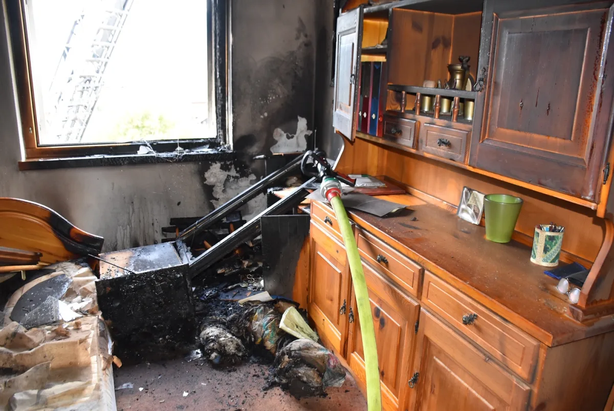 Ein Innenraum mit schweren Brandschäden, verbrannten Möbeln und einem Feuerwehrschlauch über einem Regal.
