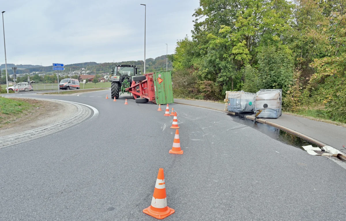 Ein Traktor mit umgekipptem Anhänger befindet sich auf einer kurvigen Straße. Verkehrshütchen markieren den Unfallbereich.