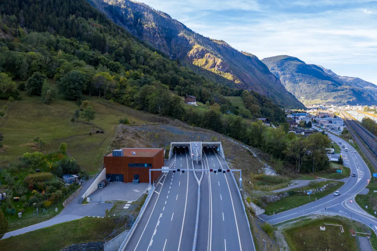Eine Autobahn führt in einen Tunnel, der in eine bergige, bewaldete Landschaft eingebettet ist. Im Hintergrund sind weitere Berge sichtbar.