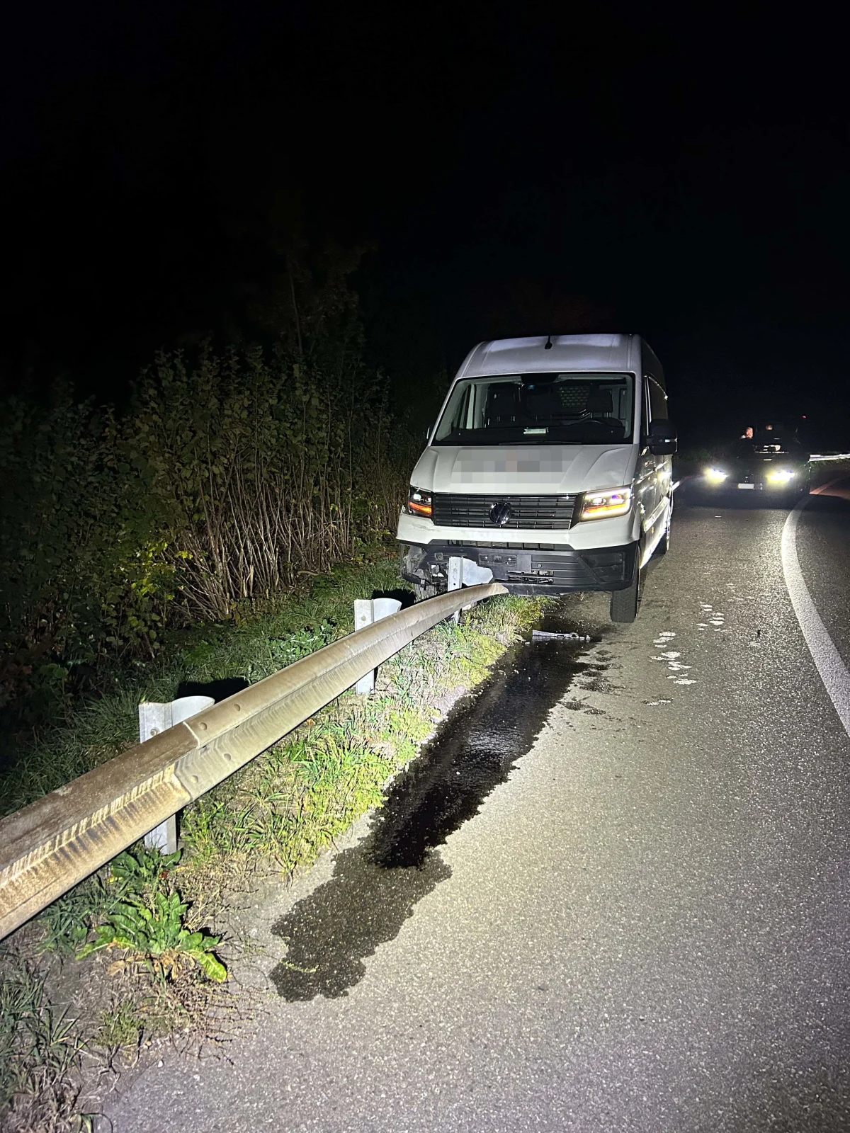 Ein weißer Lieferwagen ist nachts auf einer Landstraße zwischen einer Leitplanke und der Straße eingeklemmt. Das Fahrzeug steht schräg, und es gibt Ölflecken auf dem Asphalt. Die Szene ist dunkel und die Beleuchtung kommt teilweise vom Fahrzeug.