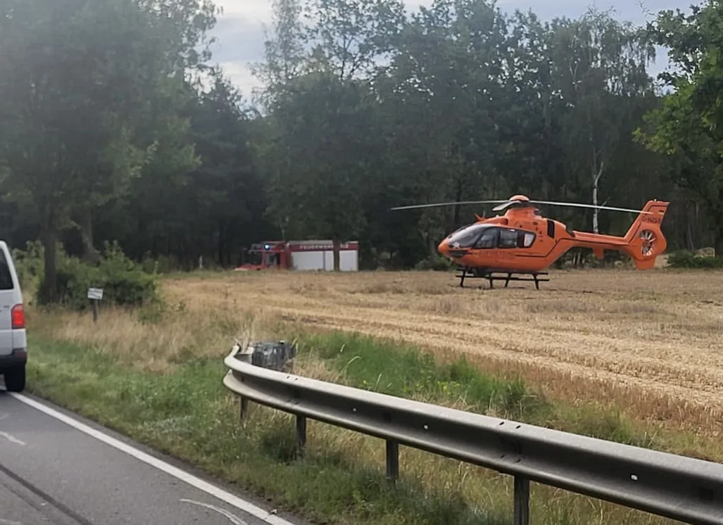 Ein Rettungshubschrauber steht auf einem Feld neben einer Landstraße, im Hintergrund ist ein Einsatzfahrzeug zu sehen.