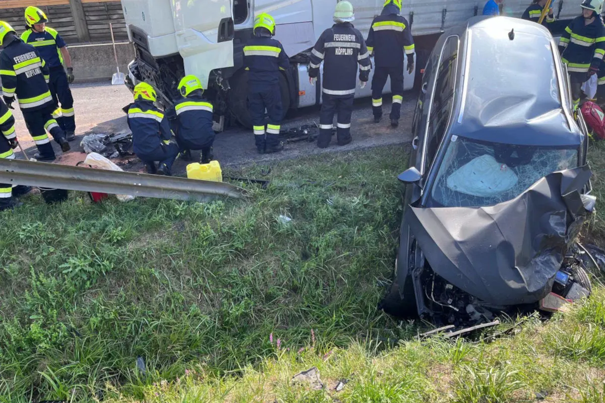 Das Bild zeigt ein schwarzes Auto mit stark beschädigter Front, das neben einer Autobahn in einem Graben steht. Im Hintergrund sichern Feuerwehrleute einen weißen Lkw, der ebenfalls in den Unfall verwickelt ist.