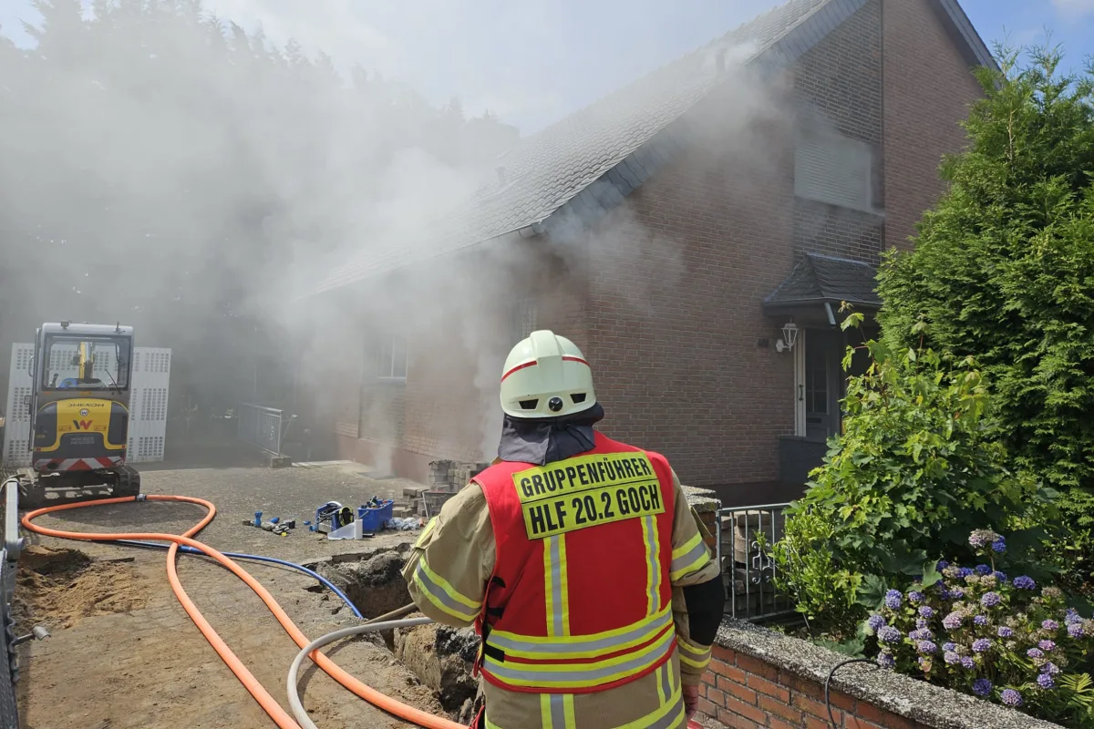 Ein Feuerwehrmann in Sicherheitsausrüstung steht vor einem Haus, aus dem Rauch strömt. Im Vordergrund sind Schläuche und ein kleiner Bagger zu sehen.