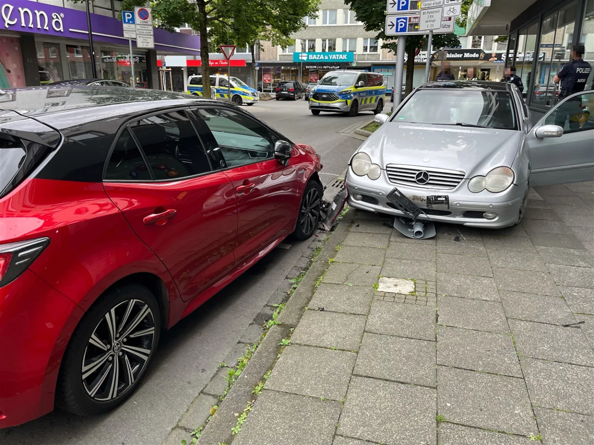 Ein silberner Mercedes steht beschädigt auf einem Gehweg und berührt einen roten Toyota. Beide Fahrzeuge weisen Unfallspuren auf.