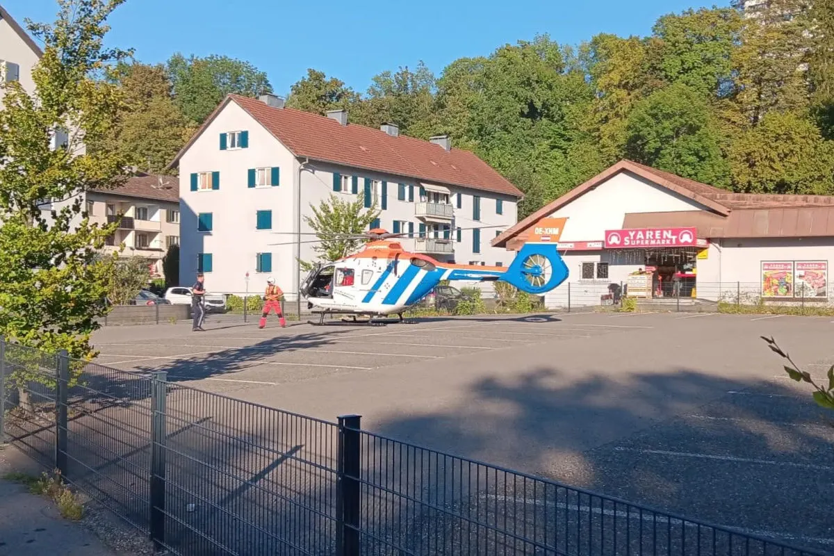 Ein Rettungshubschrauber steht auf einem städtischen Parkplatz neben mehreren Gebäuden bei klarem Himmel. Zwei Personen stehen in der Nähe.