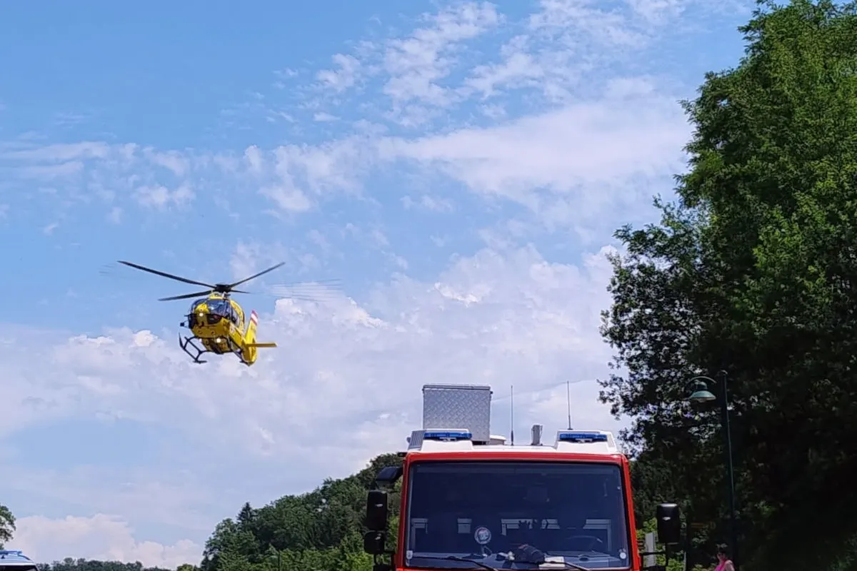 Ein gelber Rettungshubschrauber schwebt am blauen Himmel über einem rot-weißen Feuerwehrfahrzeug. Bäume sind im Hintergrund sichtbar.