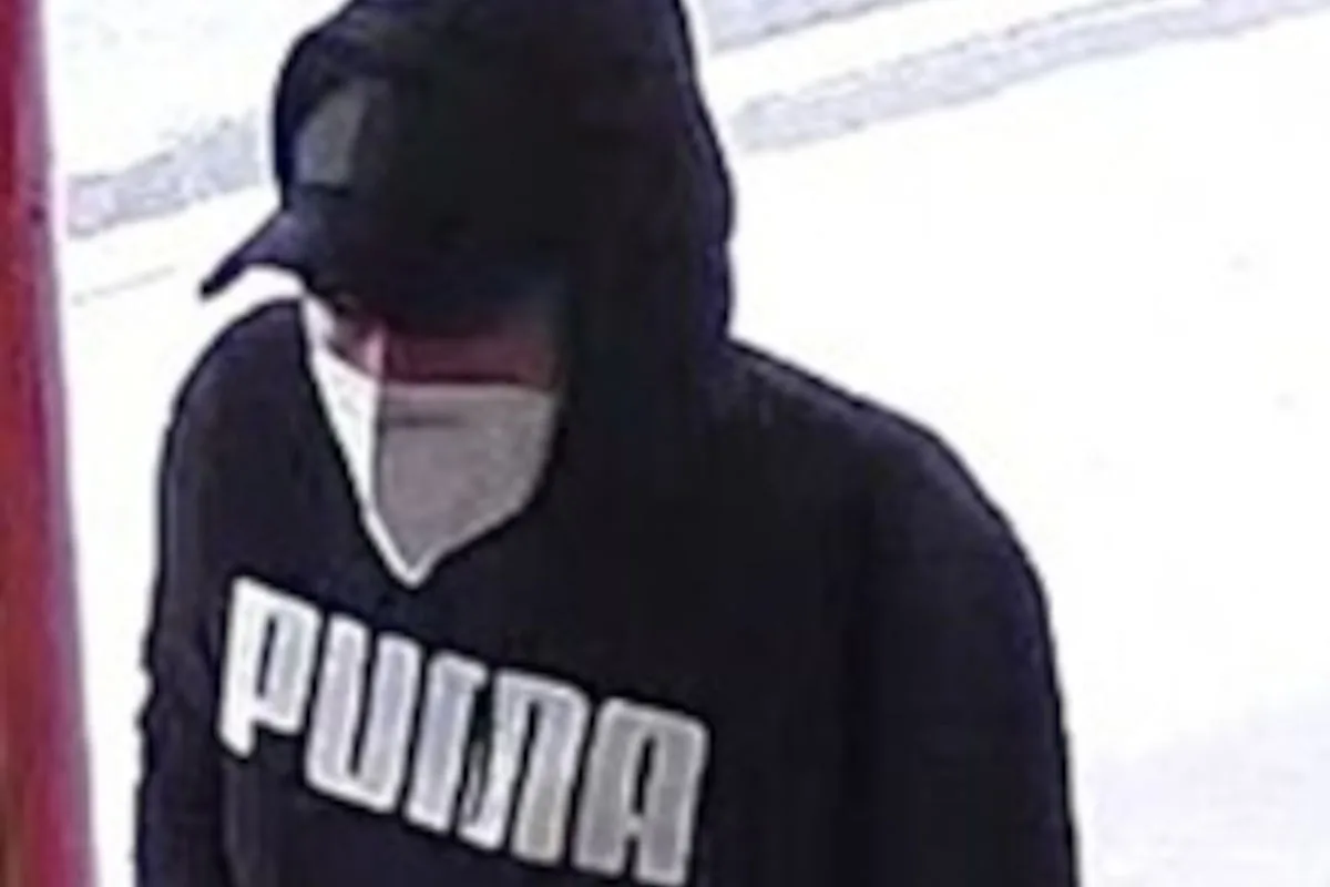 Eine Person mit schwarzer Kapuzenjacke und Baseballkappe trägt eine weiße Maske. Die Umgebung ist hell beleuchtet.