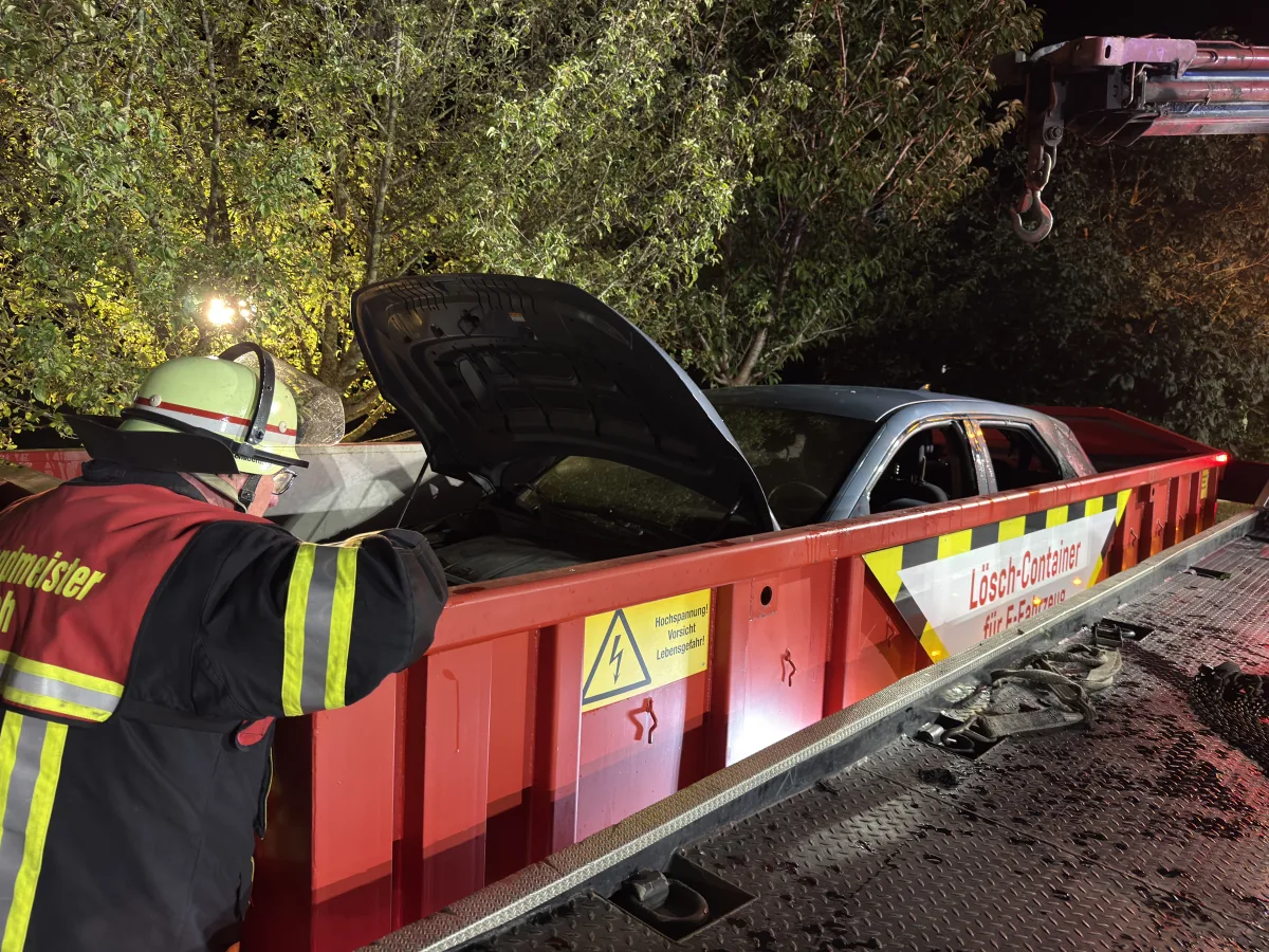 Ein Feuerwehrmann inspiziert einen PKW in einem roten Lösch-Container bei Nacht unter Bäumen.