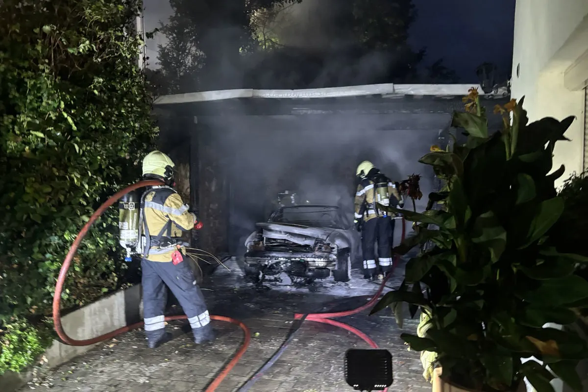 Zwei Feuerwehrleute in Schutzkleidung sind damit beschäftigt, ein brennendes Auto in einer Garage zu löschen. Rauch steigt auf.