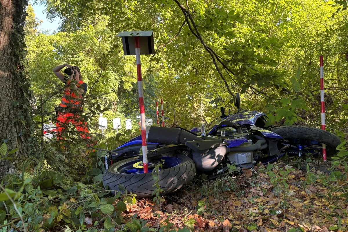 Ein blaues Motorrad liegt gestürzt am Boden im Gebüsch. Ein Mann in orangefarbener Weste fotografiert die Szene.
