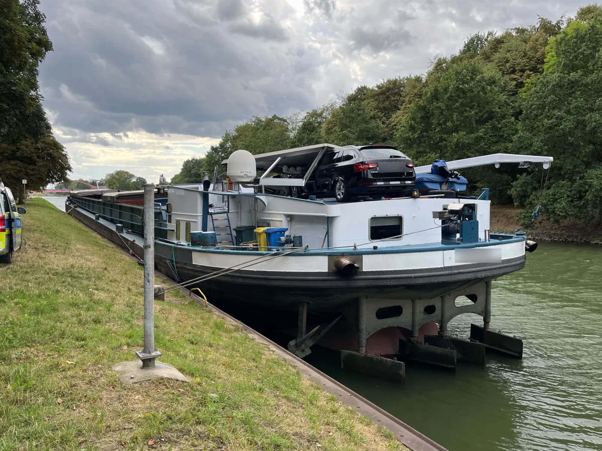 Ein schwarzes Auto befindet sich auf einem Lastkahn, der an einem Kanal entlang fährt; es ist bewölkt.