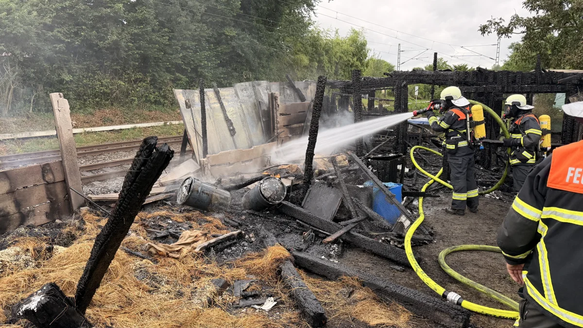 Zwei Feuerwehrleute in Schutzkleidung richten einen Wasserschlauch auf die Überreste eines abgebrannten Schuppens. Bahngleise sind im Hintergrund zu sehen.