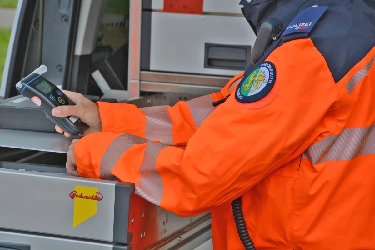 Ein Polizist in einer auffällig orangefarbenen Jacke hält ein Alkoholmessgerät neben einem Einsatzfahrzeug. Auf der Jacke ist das Emblem der Kantonspolizei zu sehen.