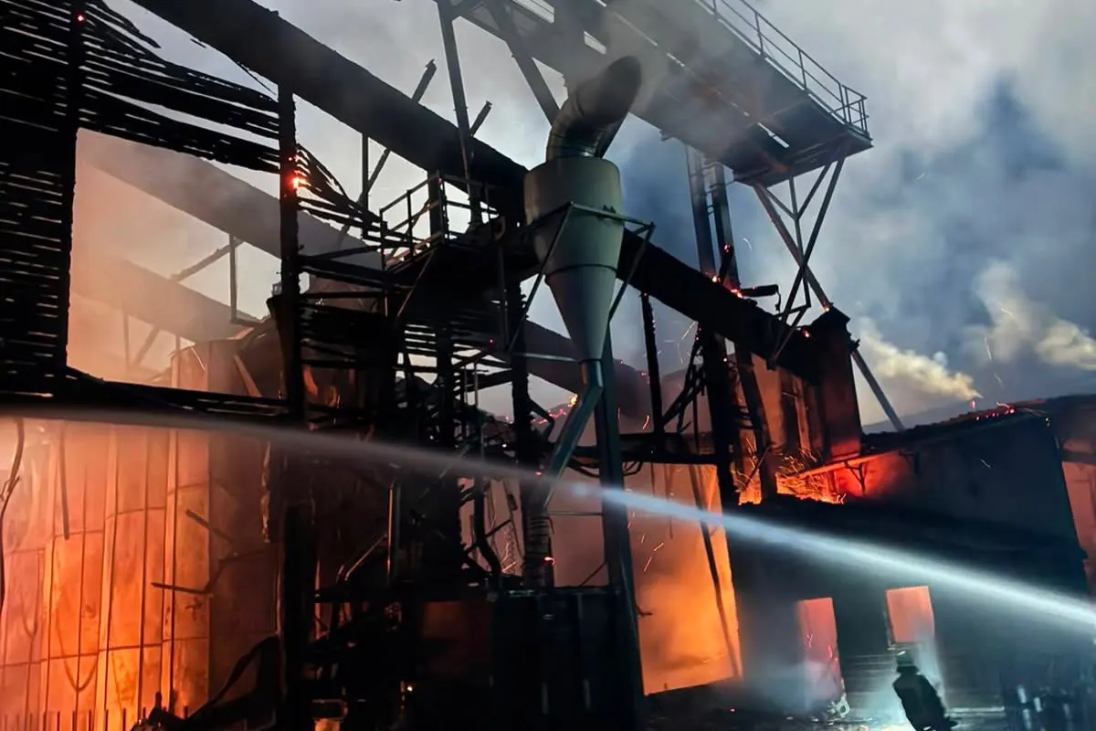 Ein Industriegebäude steht in Flammen, während die Feuerwehr mit einem Wasserschlauch das Feuer bekämpft. Funken und Rauch sind sichtbar.
