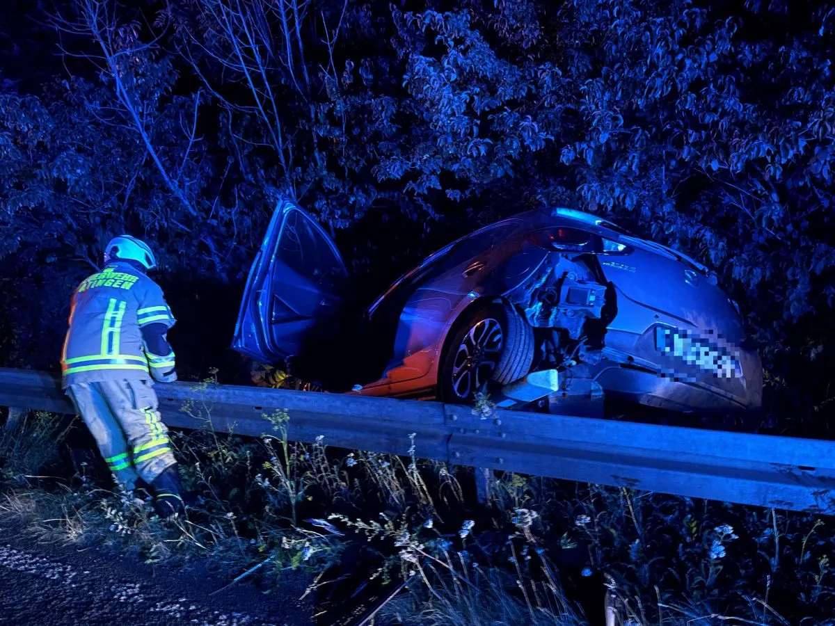 Ein Auto hat sich in einer Leitplanke verkeilt. Ein Feuerwehrmann steht daneben, die Szene ist nachts beleuchtet.