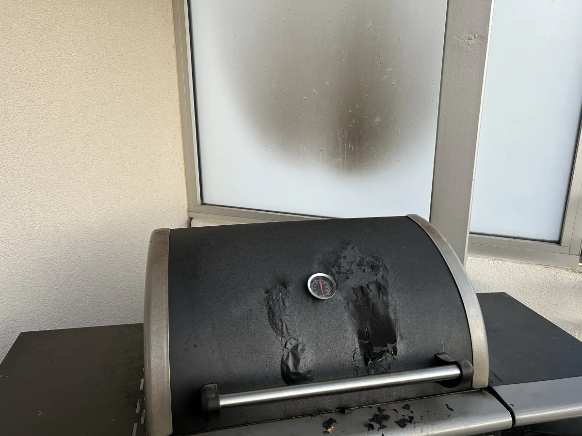 Ein dunkler Grill mit erkennbaren Brandspuren steht vor einer matten Scheibe, die durch den Rauch verfärbt ist.