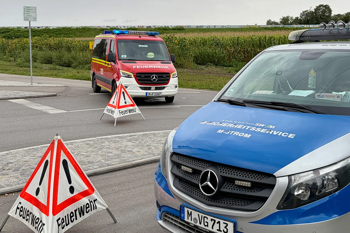 Ein rotes Feuerwehrfahrzeug steht auf einer gesperrten Straße, flankiert von einem blauen Sicherheitsfahrzeug. Warnschilder mit der Aufschrift 'Feuerwehr' sperren die Straße ab.