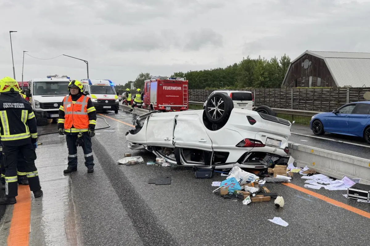 Ein weißes Auto liegt nach einem Unfall auf dem Dach auf der nassen Fahrbahn, umgeben von Feuerwehr- und Rettungsfahrzeugen.
