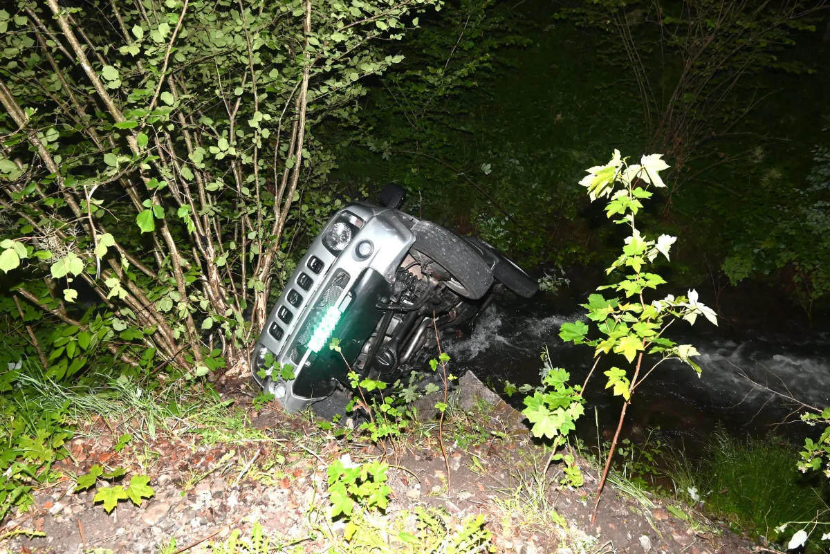 Ein silberner Jeep liegt auf seiner Seite in einem flachen Bachbett. Der Wagen befindet sich neben einem steilen Abhang, umgeben von dichtem Grün.