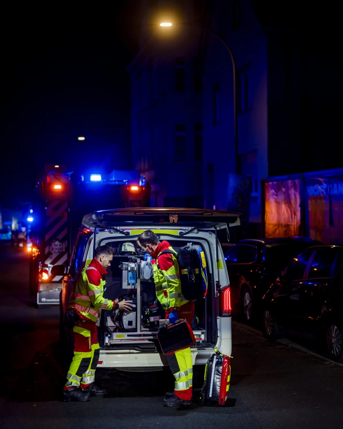 Zwei Rettungskräfte in gelb-roten Uniformen entladen Ausrüstung aus einem offenen weißen Einsatzfahrzeug. Die Szene ist nachts, beleuchtet von Straßenlaternen und den Blaulichtern weiterer Einsatzfahrzeuge.