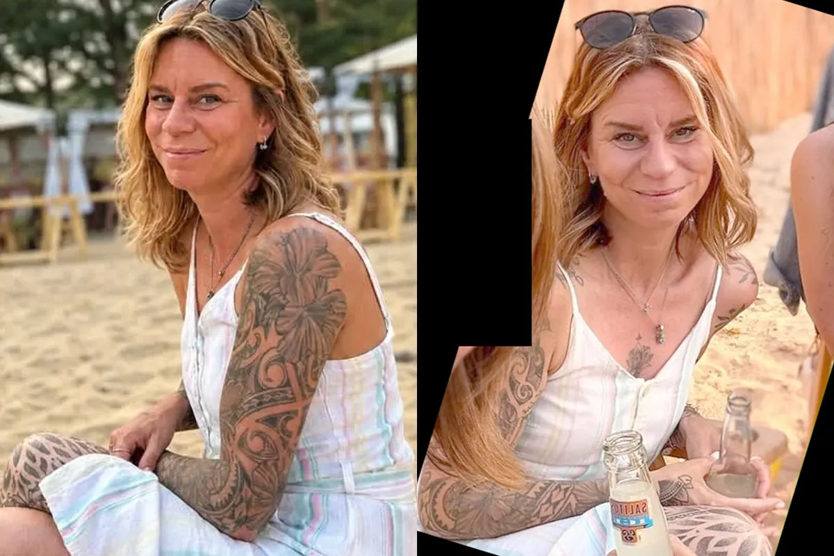 Eine Frau mit blonden Haaren und Tattoos auf den Armen sitzt am Strand und lächelt in die Kamera. Sie trägt ein ärmelloses Kleid und hat eine Sonnenbrille auf dem Kopf.