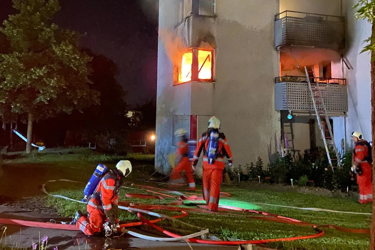 Mehrere Feuerwehrleute in Schutzanzügen löschen in der Nacht einen Brand in einem Wohnhaus mit Schläuchen. Ein Fenster im ersten Stock steht in Flammen.