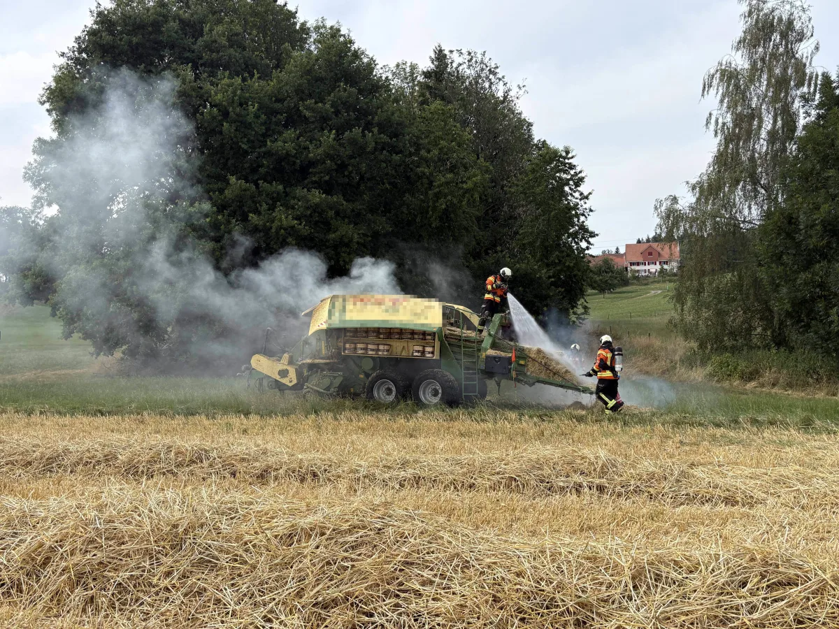Zwei Feuerwehrleute bekämpfen einen Brand an einem Mähdrescher auf einem offenen Feld. Rauch steigt auf, während im Hintergrund Bäume und ein Gebäude sichtbar sind.