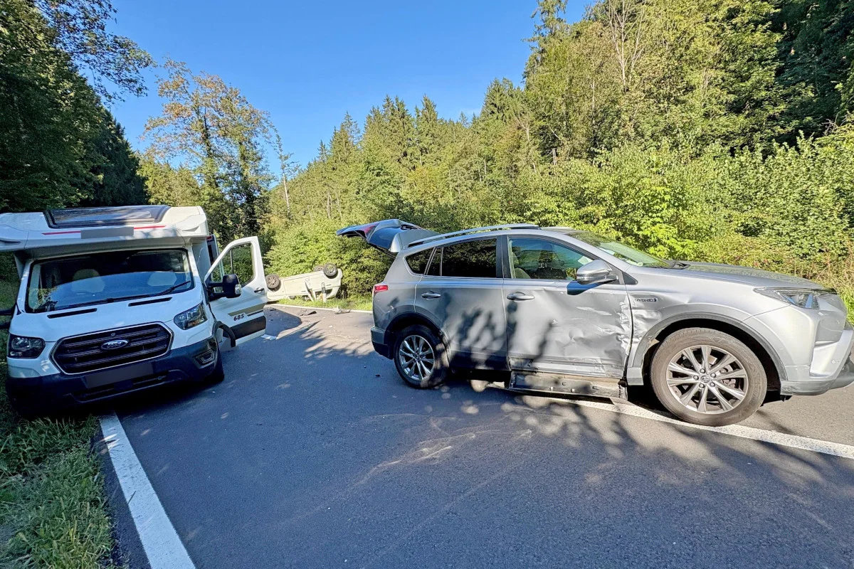 Auf einer Landstraße am Waldrand steht ein Wohnmobil, daneben ein SUV; im Hintergrund liegt ein umgestürztes Auto.