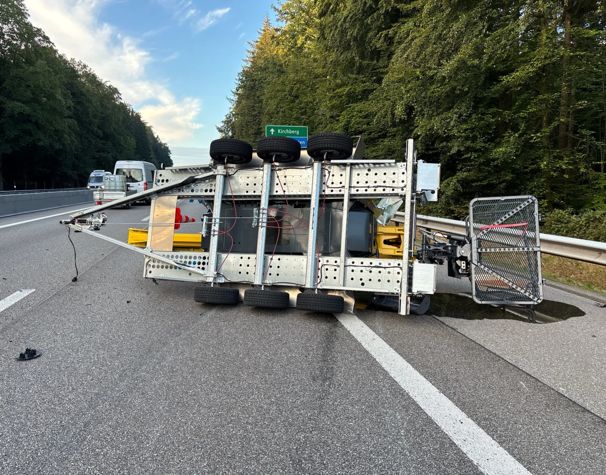 Ein umgekippter Anhänger liegt auf der Autobahn, während Autos im Hintergrund vorbeifahren. Straßenverkehrsschilder und Waldsäume sind sichtbar.