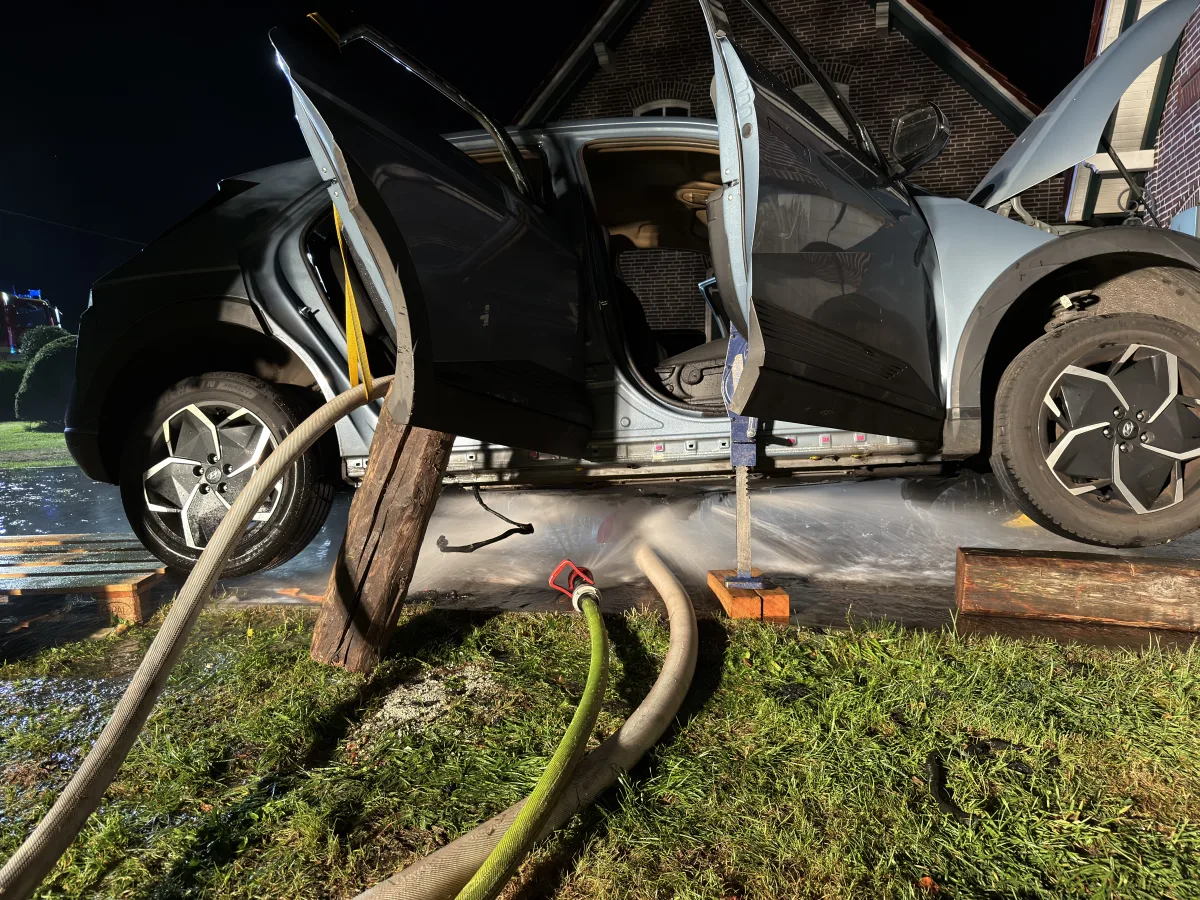 Ein Auto steht mit offenen Türen auf Holzblöcken vor einem Gebäude. Schläuche und Wasser sind im Vordergrund sichtbar.
