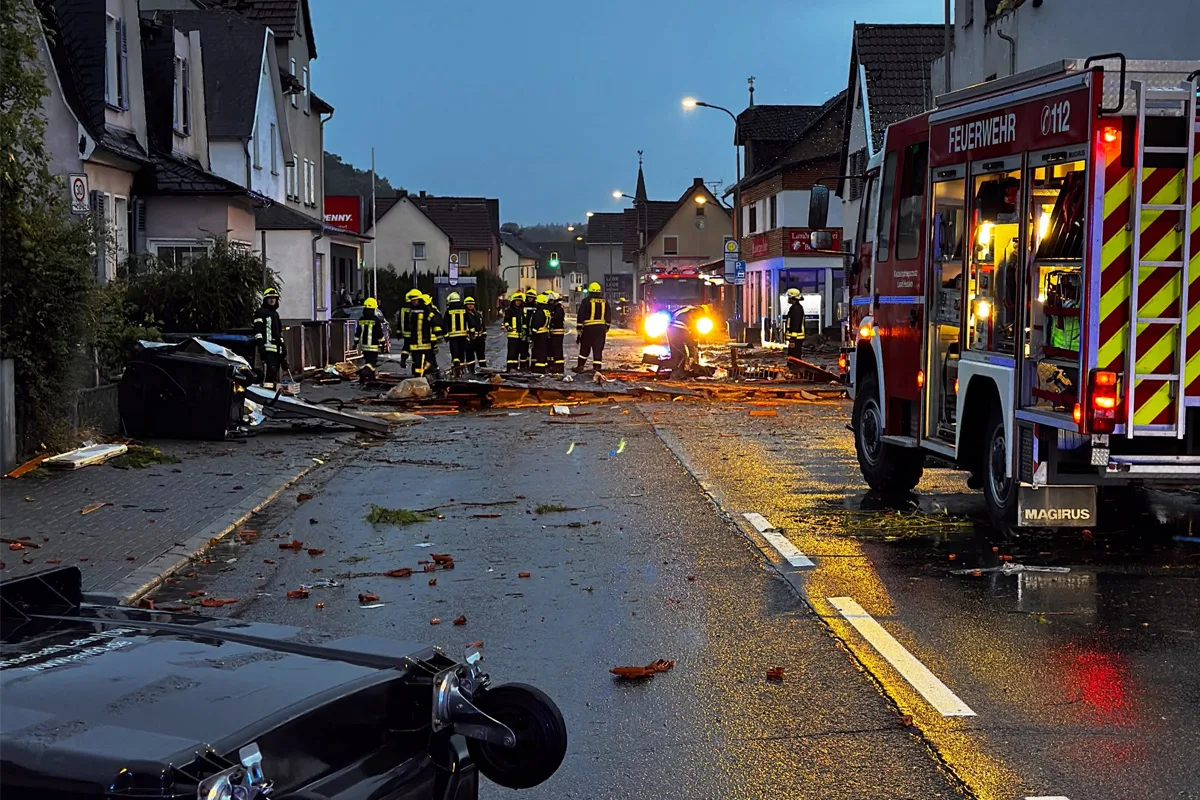 Feuerwehrleute stehen auf einer Straße und räumen Trümmer beiseite. Ein Feuerwehrfahrzeug steht rechts im Bild.