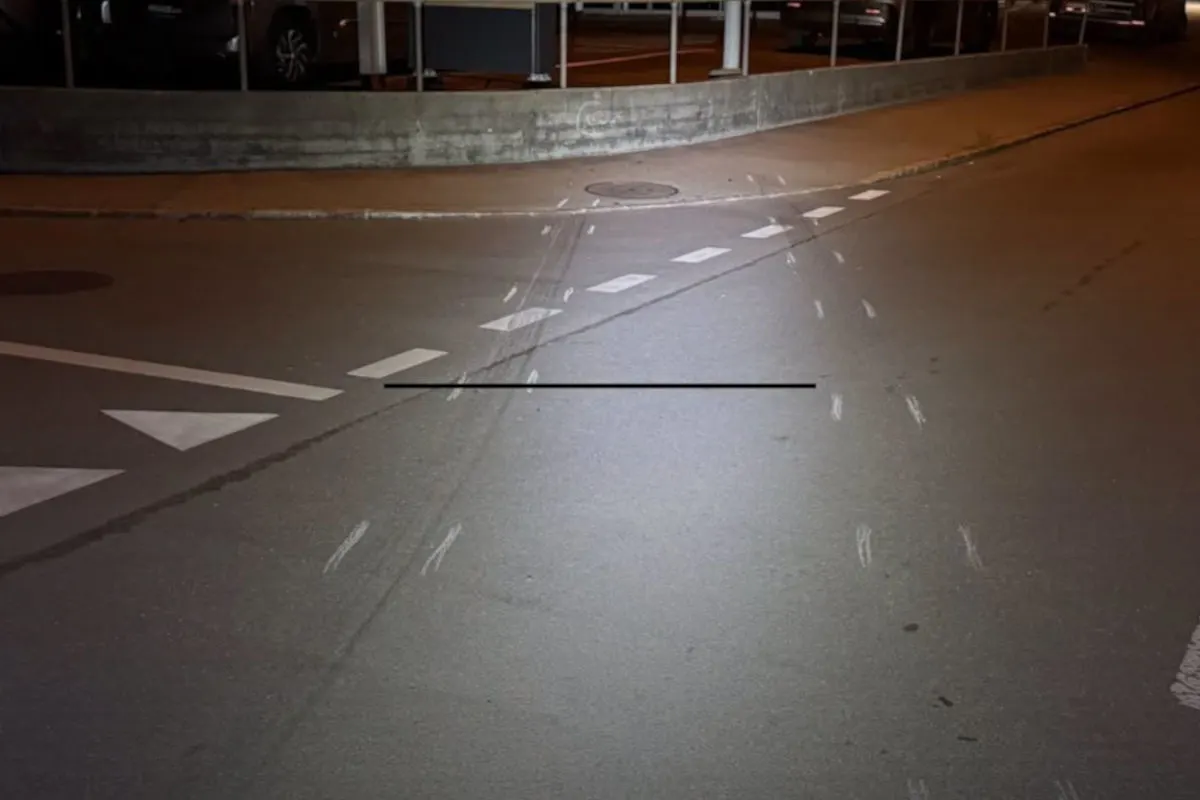Auf einer dunklen Straße sind deutliche Reifenspuren an einer Kreuzung zu sehen. Die Szenerie ist von einem Bordstein und mehreren parkenden Autos gesäumt.