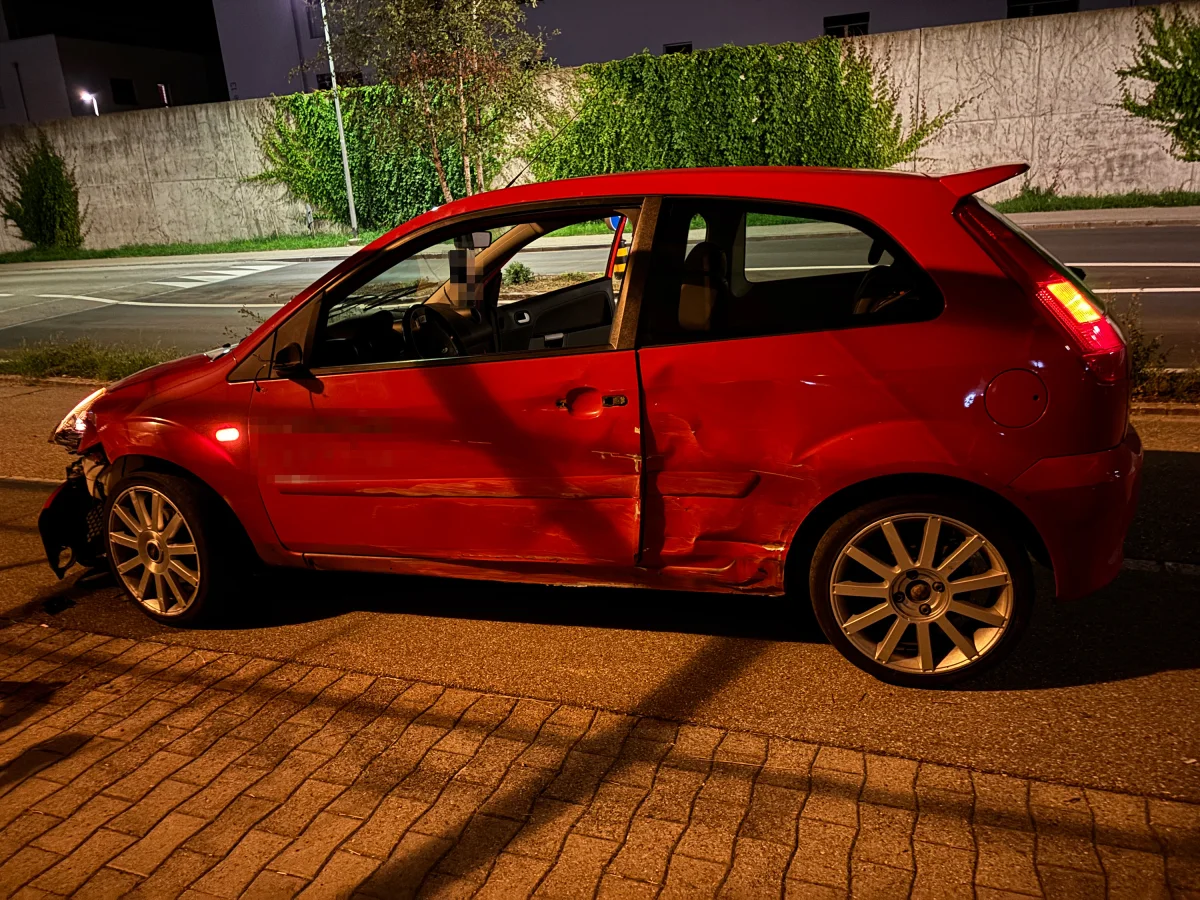 Ein rotes Auto zeigt deutliche Unfallspuren auf der Beifahrerseite. Es steht bei Nacht am Straßenrand.