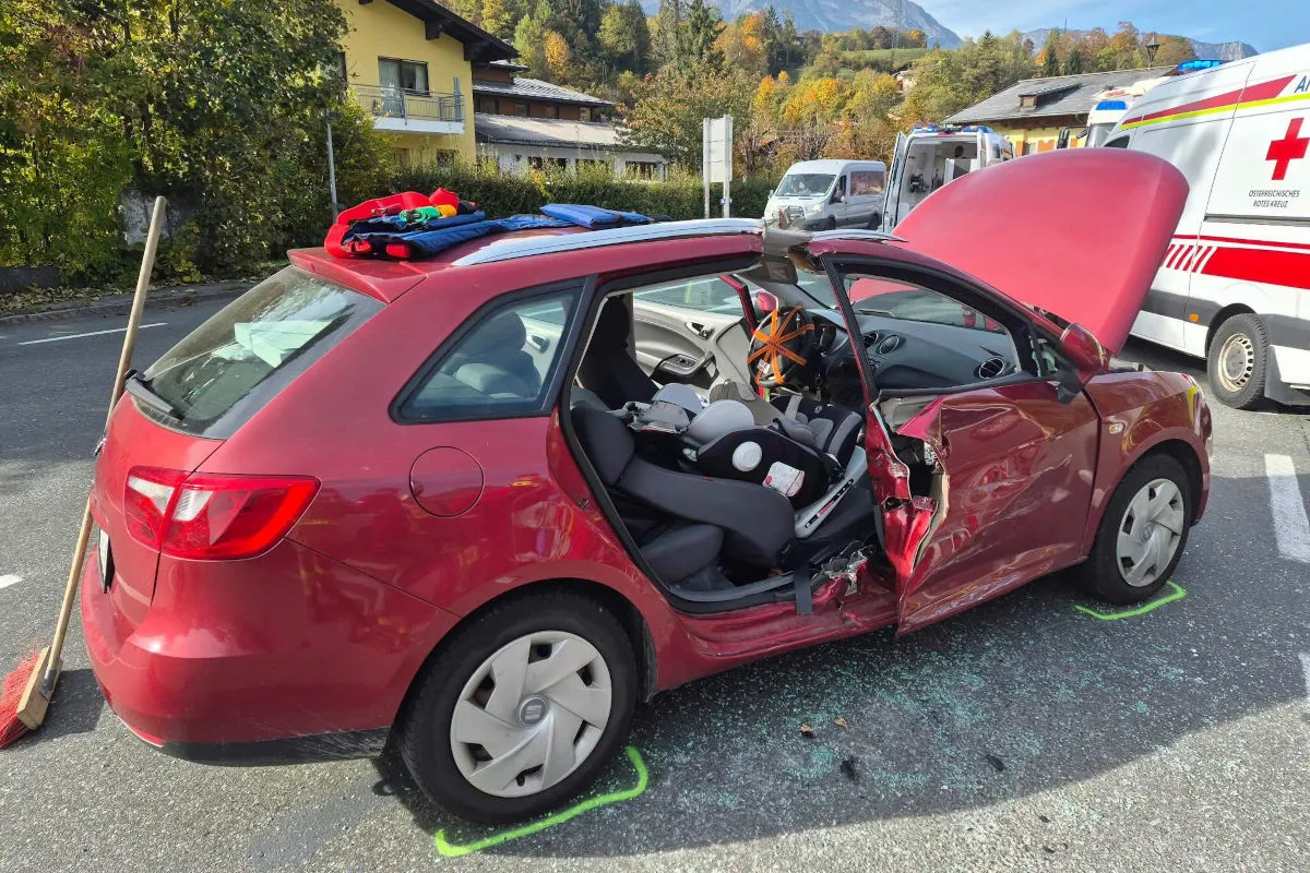 Ein rotes Auto mit offenem Kofferraum und beschädigten Türen steht auf einer Straße. Im Hintergrund ist ein Rettungswagen sichtbar.