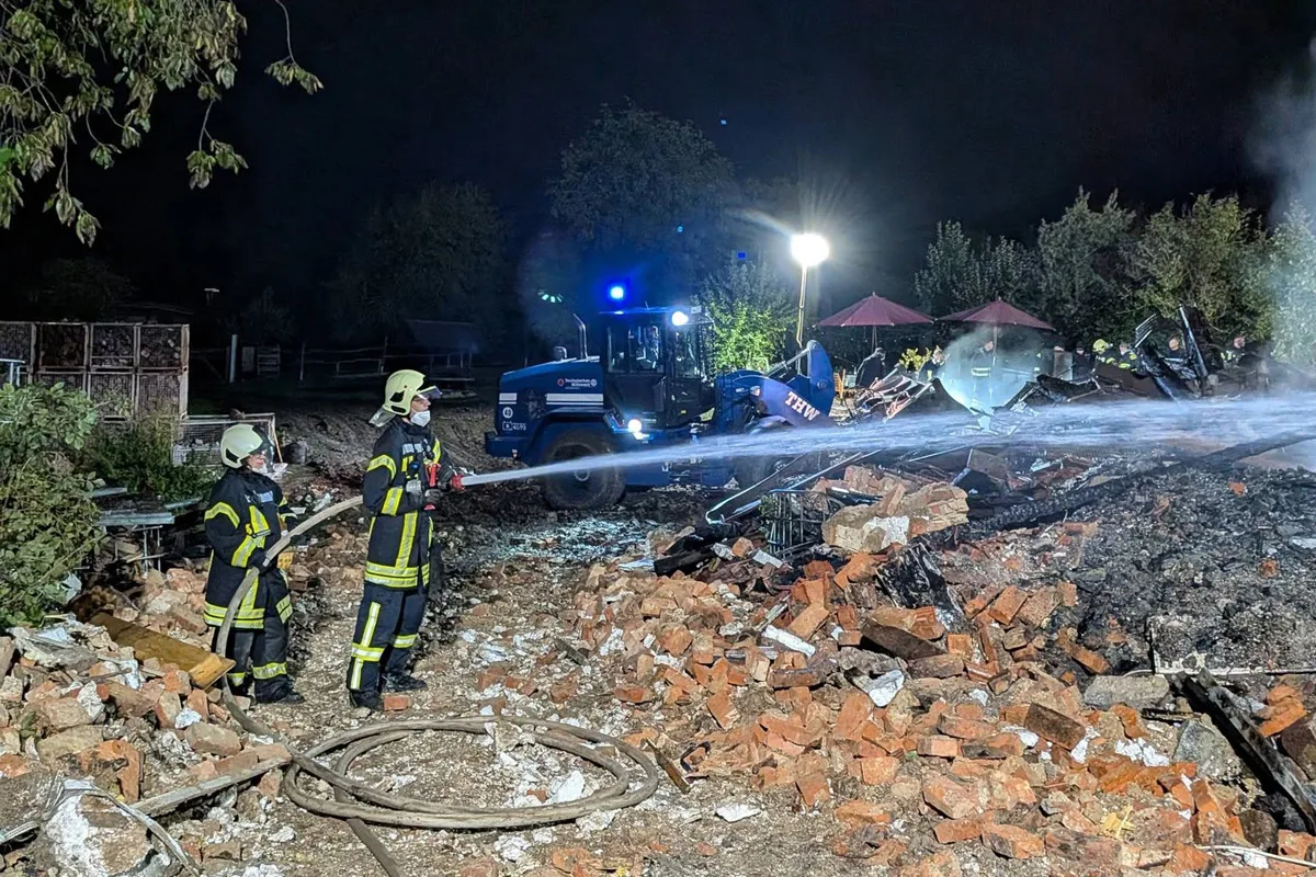 Zwei Feuerwehrleute in Schutzkleidung löschen nachts ein Feuer in einem Gebiet mit Ziegeltrümmern und heruntergebrannten Holzresten. Ein Fahrzeug des Technischen Hilfswerks ist auch vor Ort.