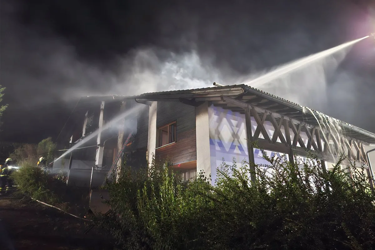 In der Nacht löschen Feuerwehrleute ein Feuer an einem zweistöckigen Haus. Wasserstrahlen richten sich auf das Dach, Rauch steigt auf.