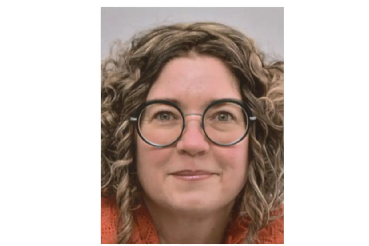 Eine Frau mit lockigem Haar und Brille blickt frontal in die Kamera. Sie trägt einen orangen Pullover.