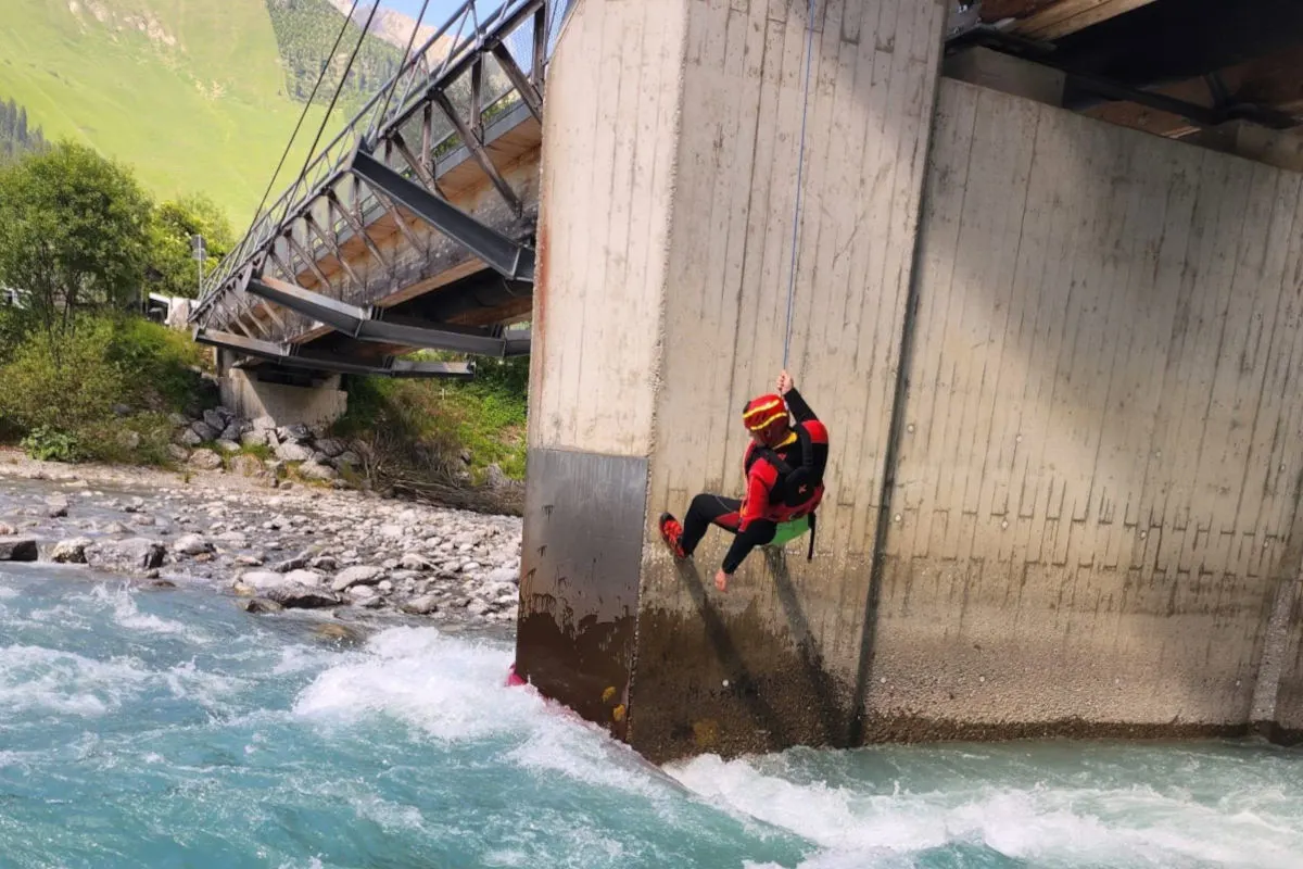 Eine Person in roter Rettungsausrüstung seilt sich an einem Brückenpfeiler ab, während unten ein Fluss fließt. Die Umgebung ist grün und bergig.