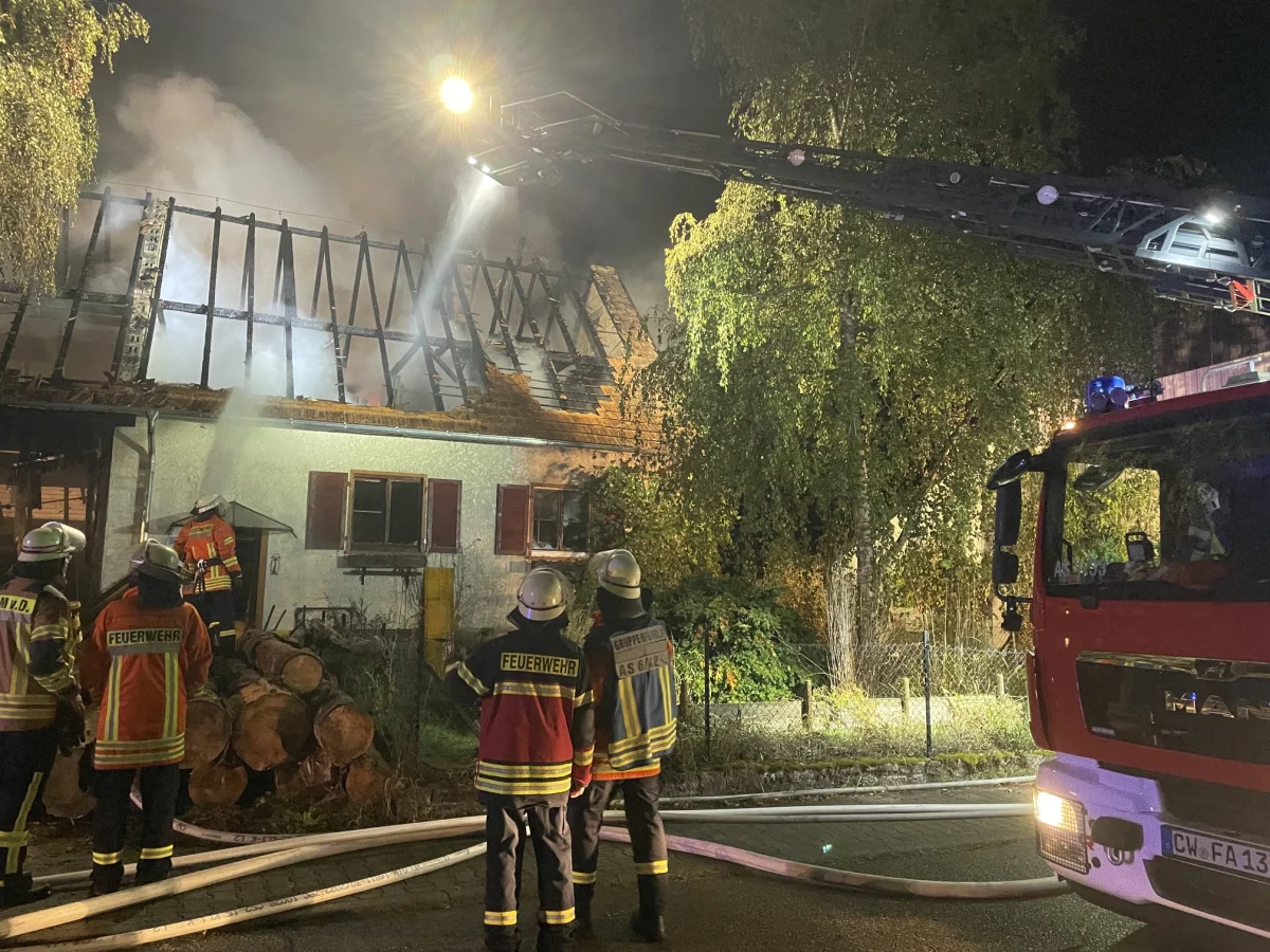 Feuerwehrkräfte arbeiten nachts an einem brennenden Haus mit beschädigtem Dachstuhl. Ein Feuerwehrfahrzeug ist im Vordergrund.