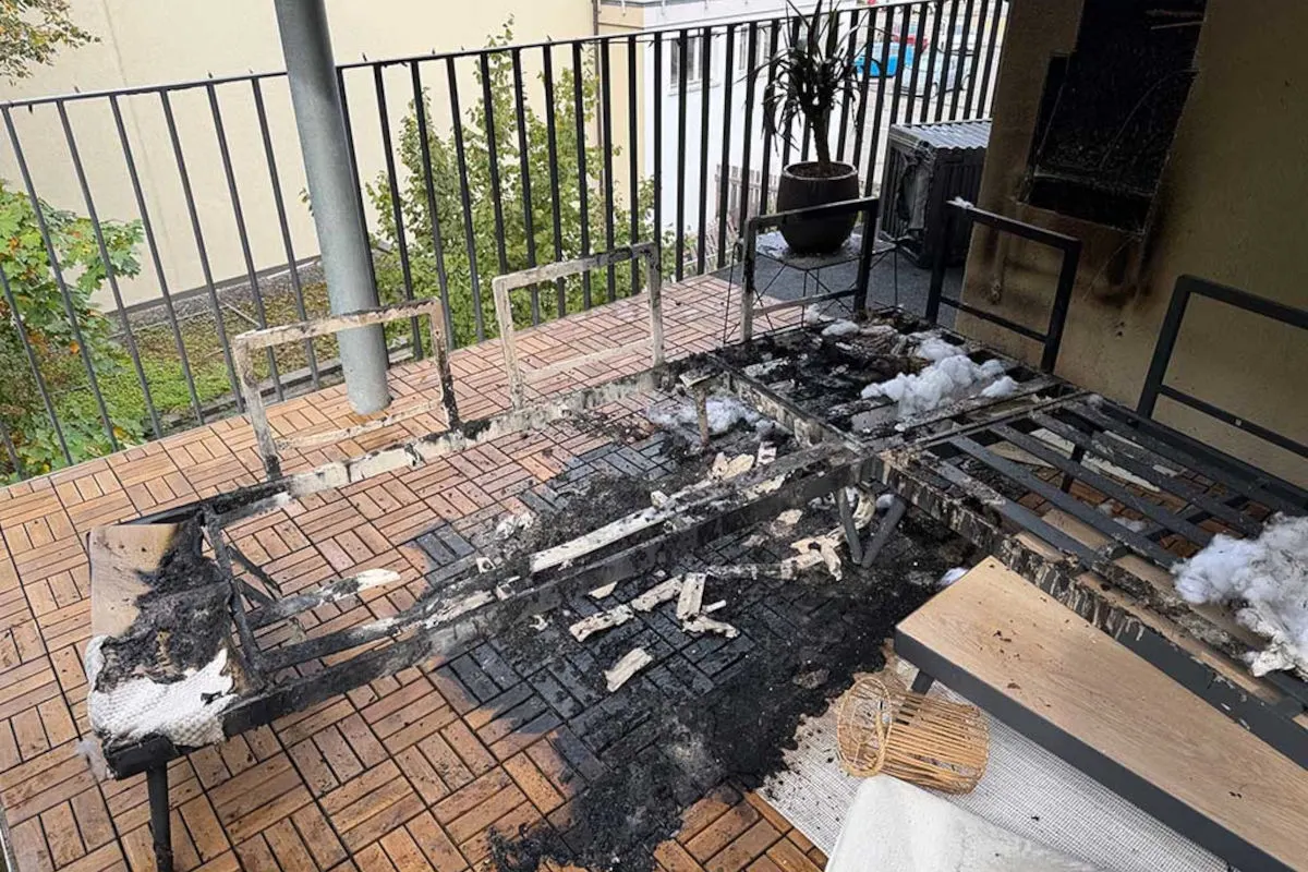 Ein Balkon zeigt die verkohlten Reste eines Metallgestells auf einem Holzboden. Im Hintergrund ist eine Balkonpflanze sichtbar.